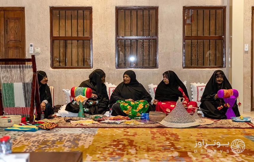 پنج زن عرب با لباس محلی، چارقد مشکی و روبنده درحال درست کردن صنایع دستی هستند.